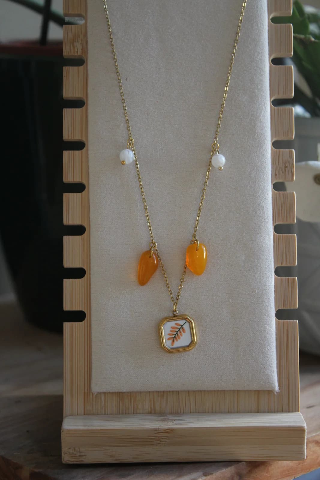 Collier doré à pampilles orange et pendentif végétal présenté sur un buste en bois.