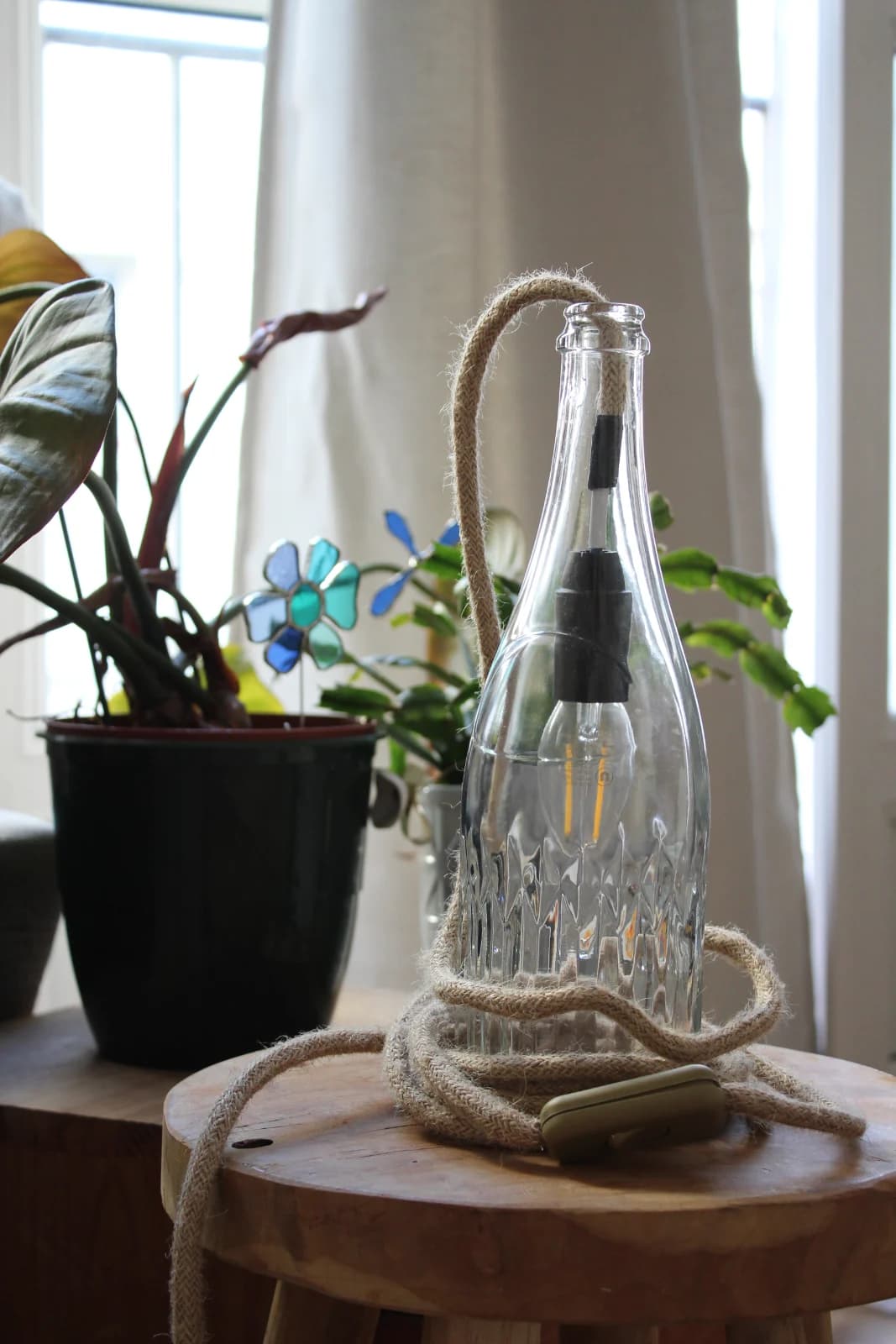 Lampe en bouteille de verre transparent éteinte, posée sur un tabouret en bois avec son câble en corde.