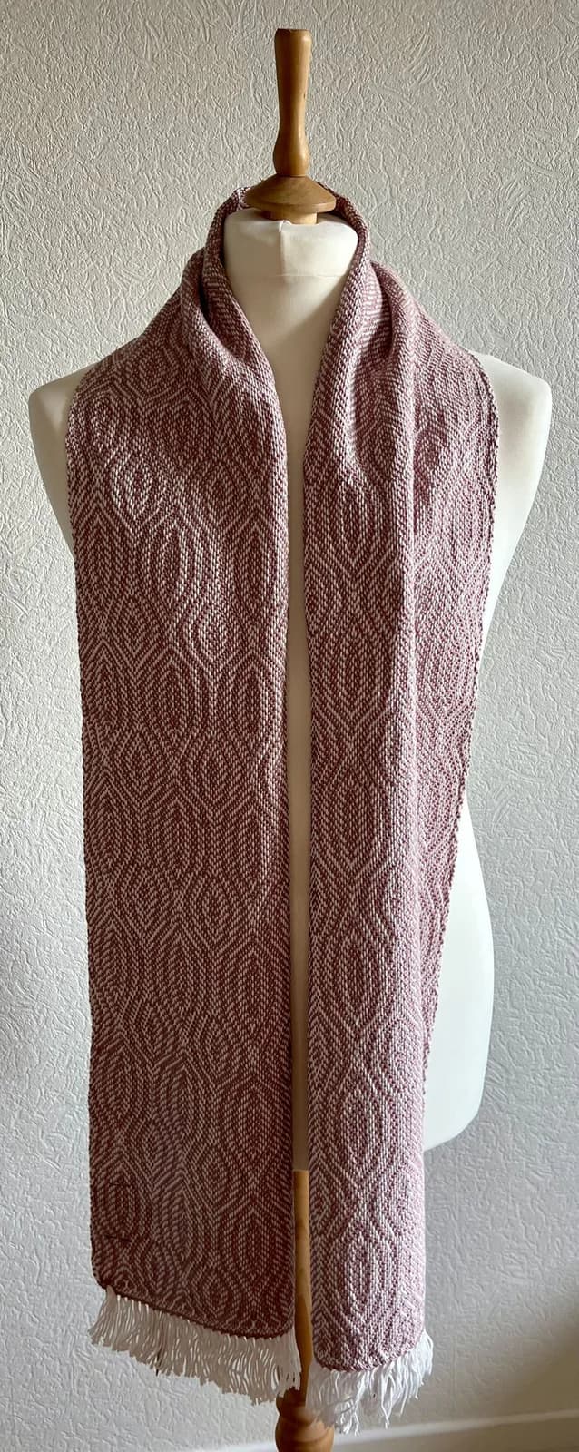 Écharpe tissée vieux rose à motifs géométriques et franges blanches sur un buste de mannequin.