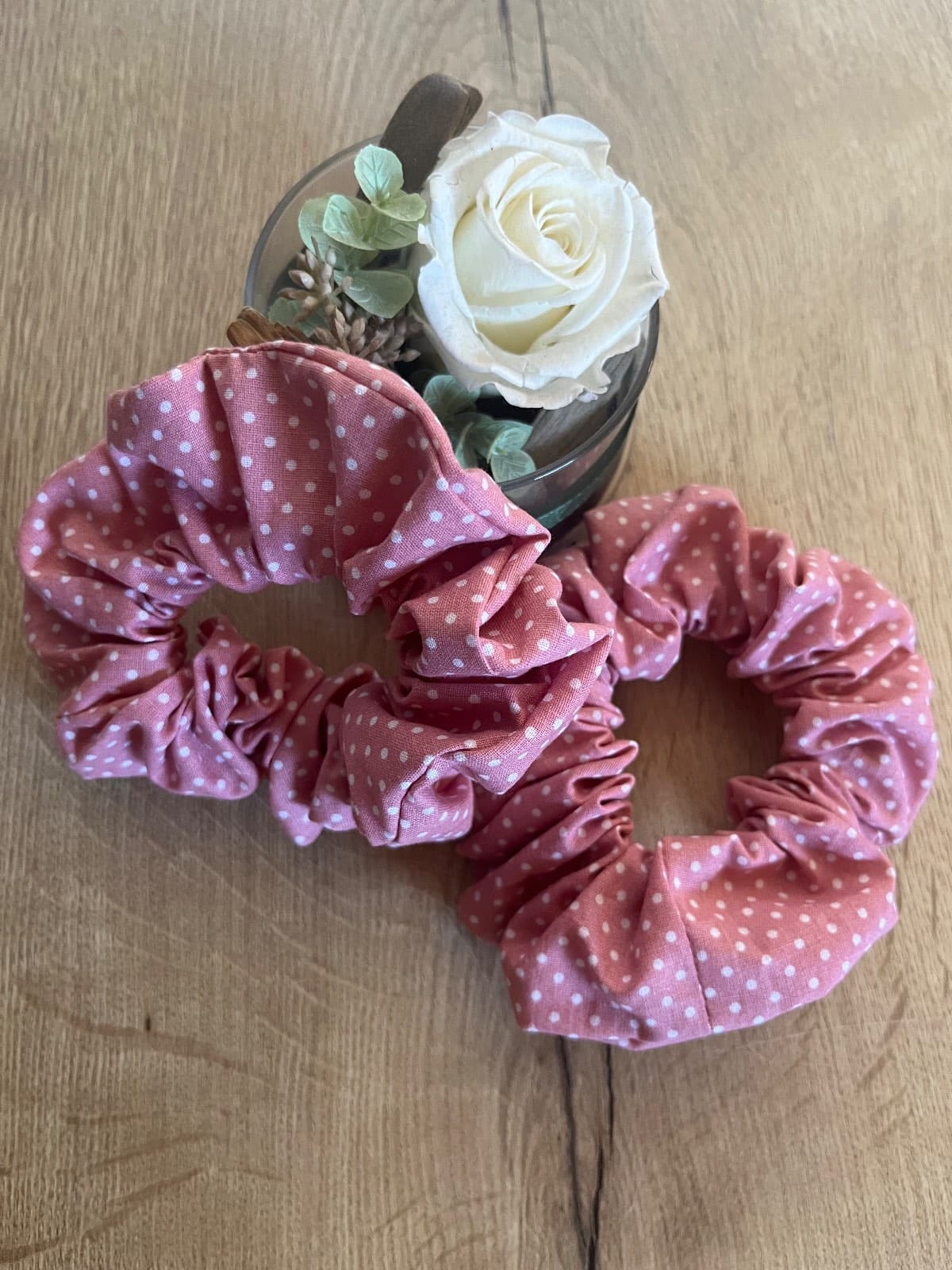 Deux chouchous froncés en tissu rose à pois blancs posés sur une table en bois à côté d'une rose.