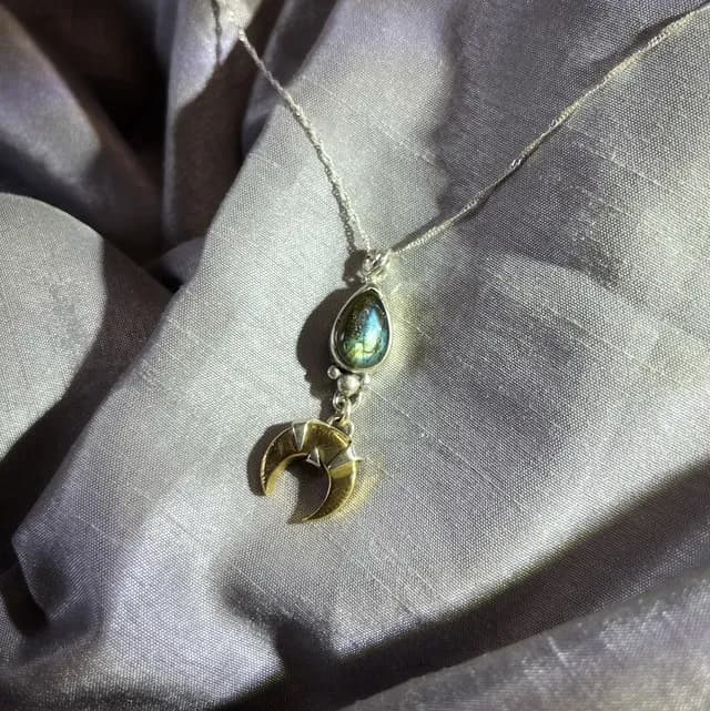 Pendentif Lunae Lumen en argent et croissant de laiton avec goutte de labradorite sur tissu gris satiné.