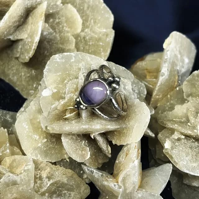 Bague en argent ornée d'un cabochon violet ovale, posée sur une rose des sables.