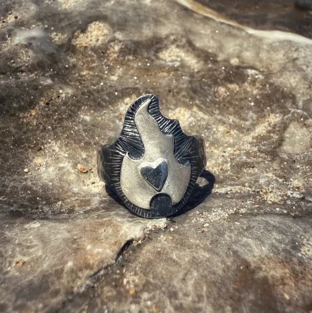 Bague argentée en forme de flamme avec relief texturé et cœur central, posée sur une roche.