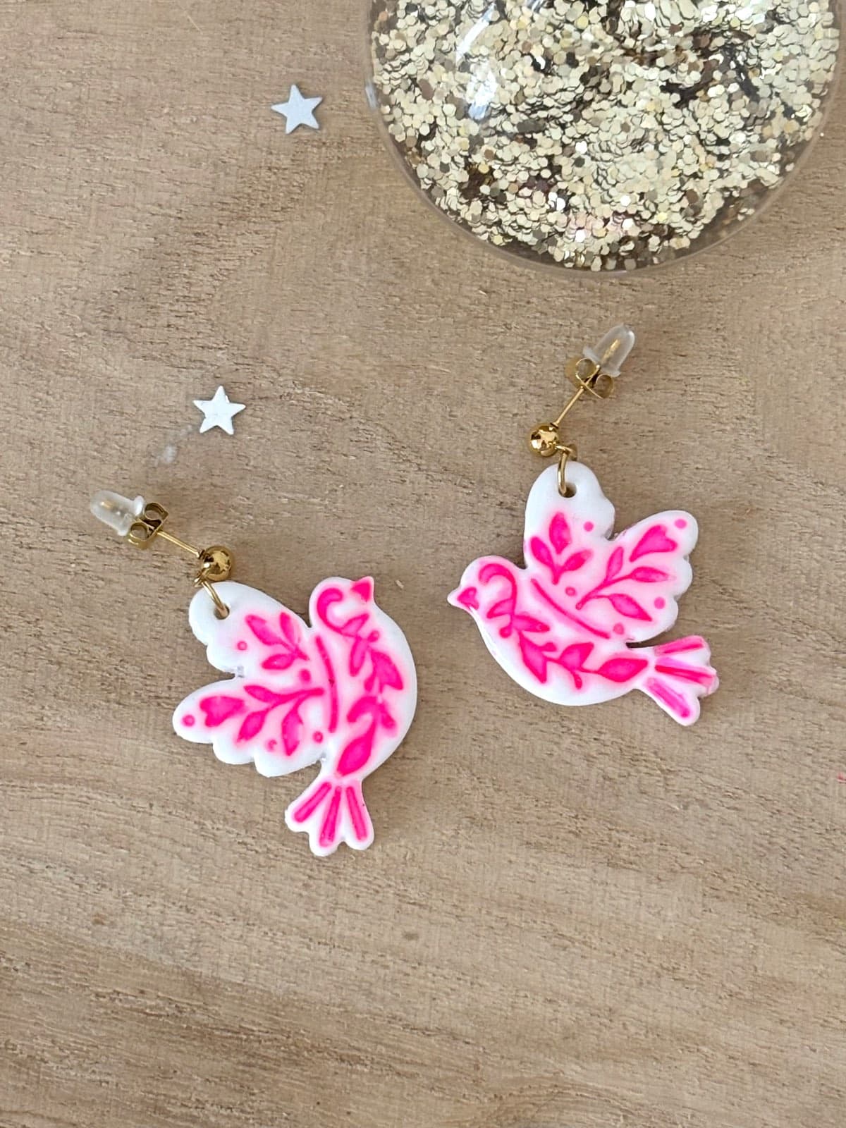 Paire de boucles d'oreilles oiseaux blancs ornés de motifs végétaux rose fluo, posée sur bois.