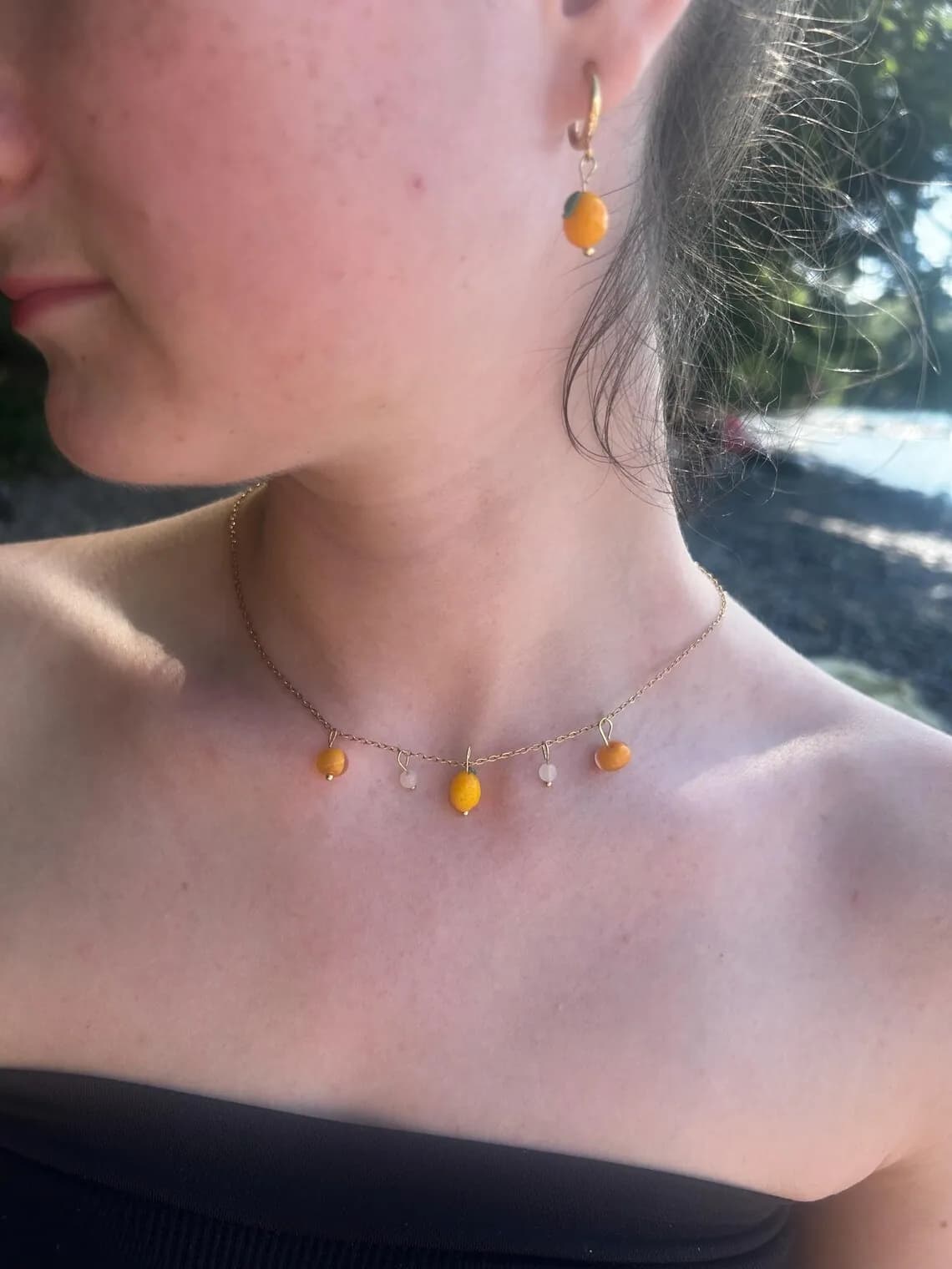 Collier doré orné de cinq charms citrons jaunes et orangés présenté sur un buste face à la mer