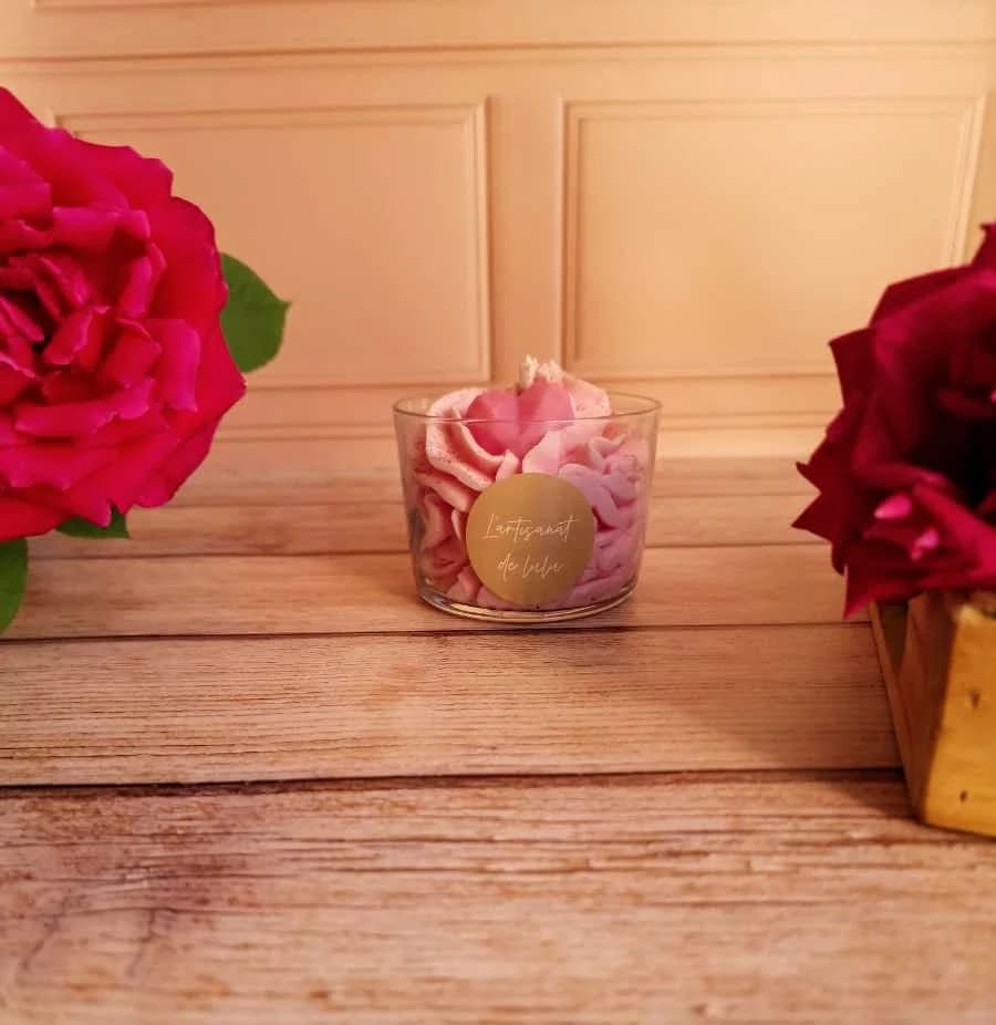 Bougie en verre avec cire rose façon chantilly et petit cœur, posée sur une table en bois entre deux roses.