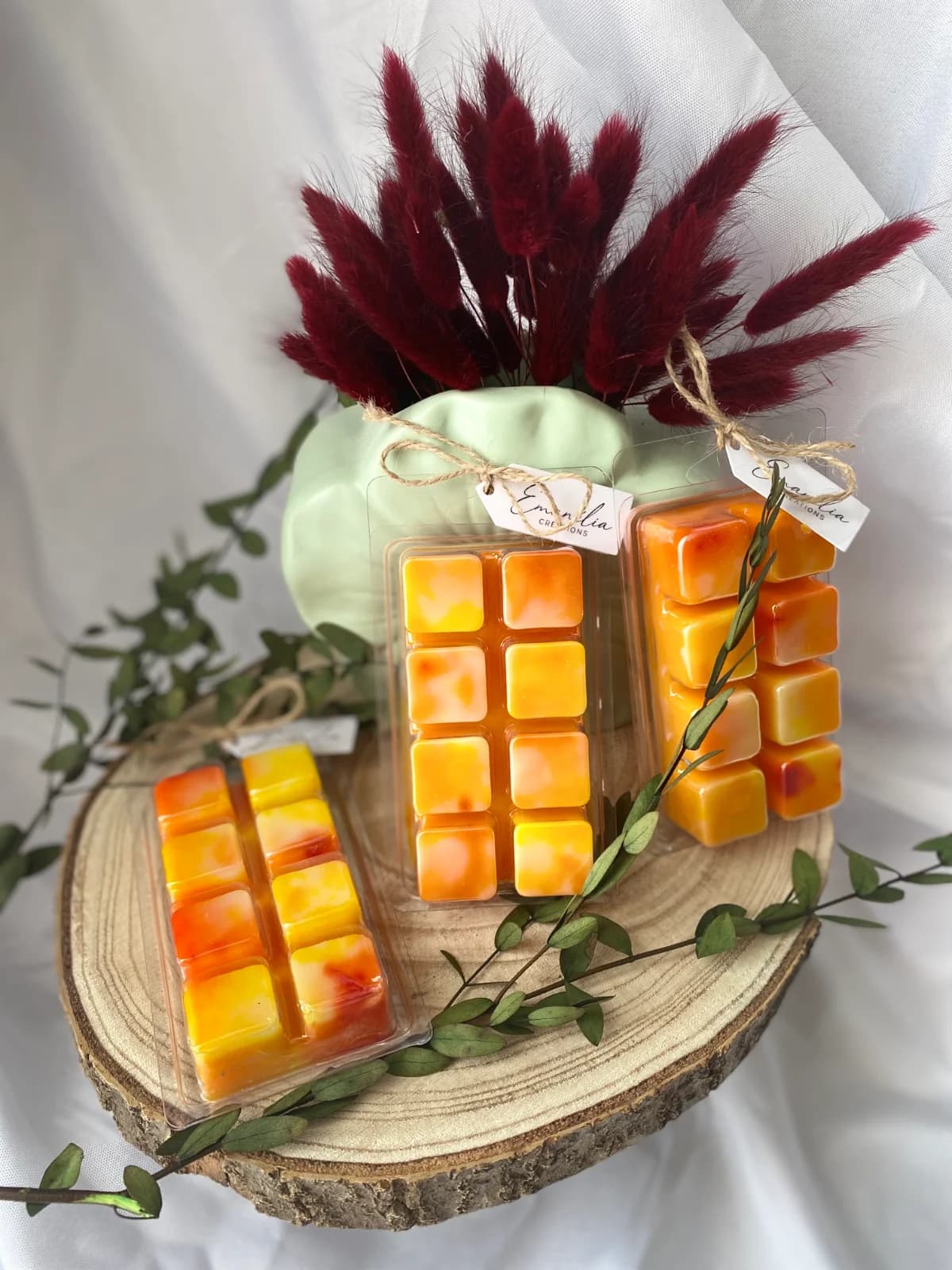 Clamshells de cire parfumée orange et jaune présentés sur un rondin de bois décoré de végétaux.